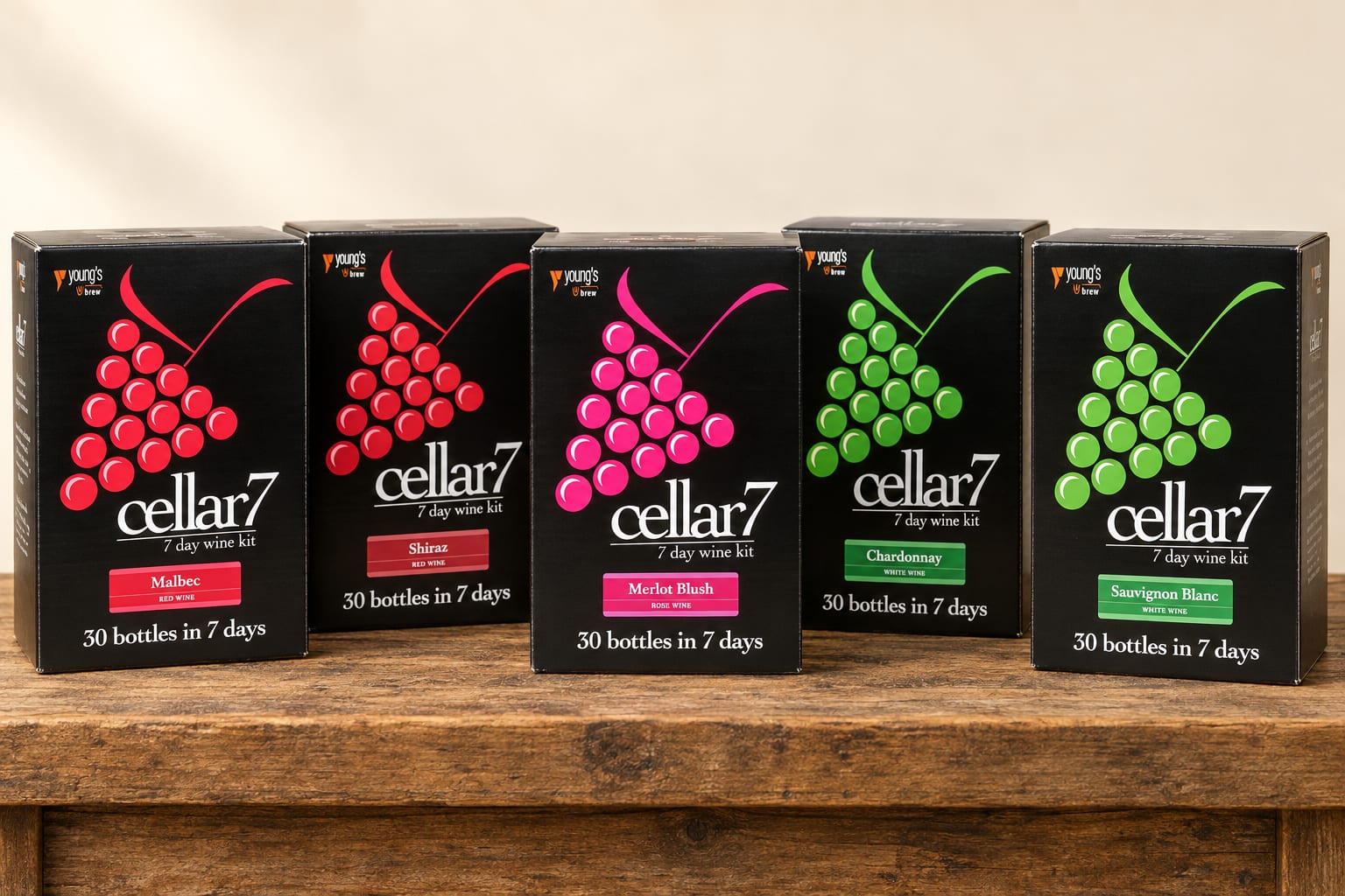 Five Cellar 7 wine kit boxes standing on a wooden bench: Malbec, Shiraz, Merlot Blush, Chardonnay and Sauvignon Blanc, each a black box with a coloured bunch of grapes on the front.